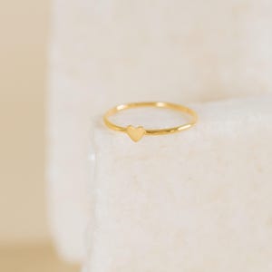 Puede incluir: Un delicado anillo dorado con un pequeño dije en forma de corazón. El anillo tiene una banda delgada con un acabado pulido, y el corazón está centrado en la parte delantera. El anillo se muestra sobre una superficie blanca y texturizada.