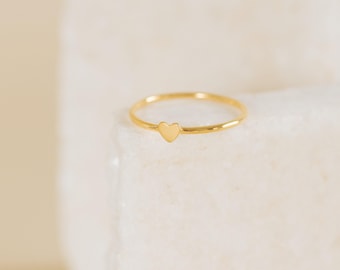 Op de afbeelding: Een delicate gouden ring met een kleine hartvormige bedel. De ring heeft een dunne band met een gepolijste afwerking en het hart is in het midden geplaatst. De ring wordt gepresenteerd op een wit, getextureerd oppervlak.