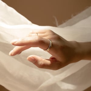 May include: A hand wearing two gold rings and one silver ring. The rings are simple bands, with a polished finish. The hand is in front of a white fabric, with a soft, natural light illuminating the scene.