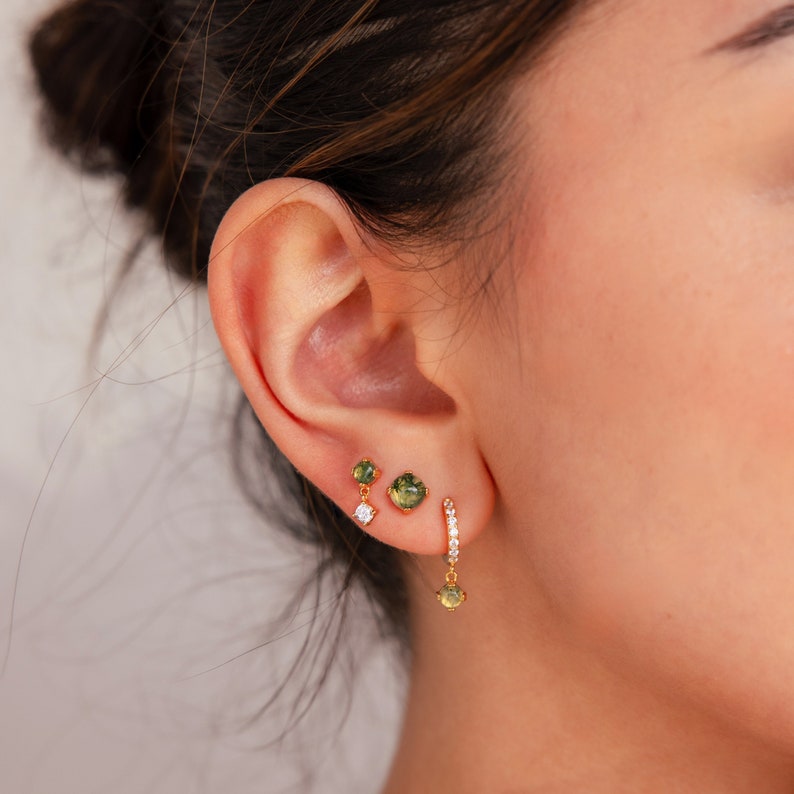 May include: A close-up of a person's ear with three gold earrings. The earrings feature green and clear gemstones.