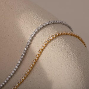 May include: Close-up of two tennis necklaces, one silver and one gold. Both necklaces are adorned with numerous small, sparkling stones, creating a continuous line of brilliance. The necklaces are displayed against a neutral, textured background.