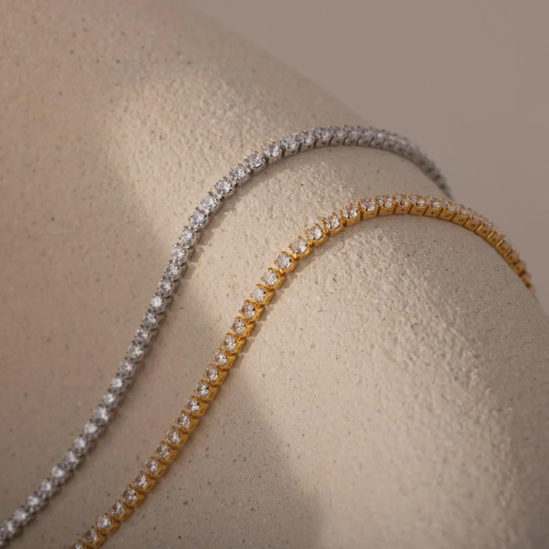 May include: Close-up of two tennis necklaces, one silver and one gold. Both necklaces are adorned with numerous small, sparkling stones, creating a continuous line of brilliance. The necklaces are displayed against a neutral, textured background.