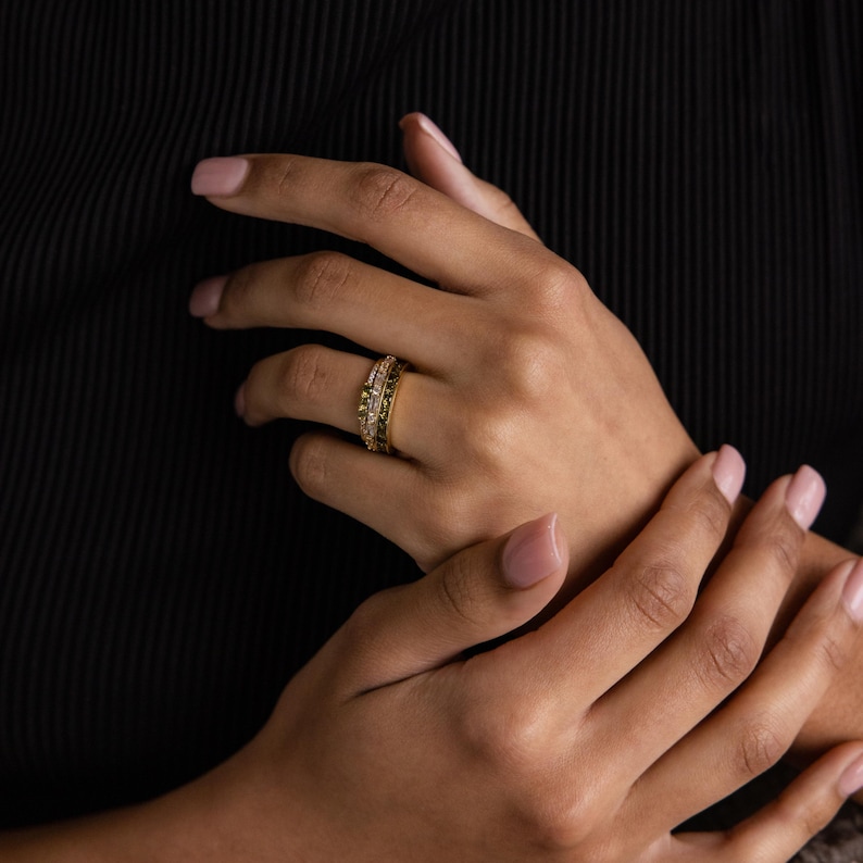 May include: Two gold rings with intricate designs are displayed on a person's hand. One ring features a row of small, clear stones, while the other has a textured, possibly floral, pattern. The background is a dark, solid color.