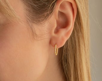 May include: Close-up of a gold hoop earring with a textured pattern. The earring is worn on an ear, with blonde hair visible. The earring is a small, circular design.