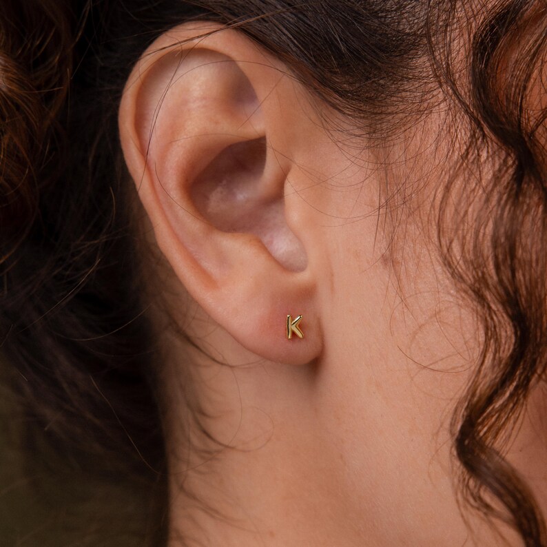 Close-up of a gold letter K stud earring worn on the ear, styled with soft curls and warm natural lighting.