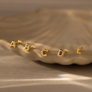 Gold initial stud earrings displaying the letters A, B, C, D, and E, arranged along the edge of a textured dish under warm, soft lighting.