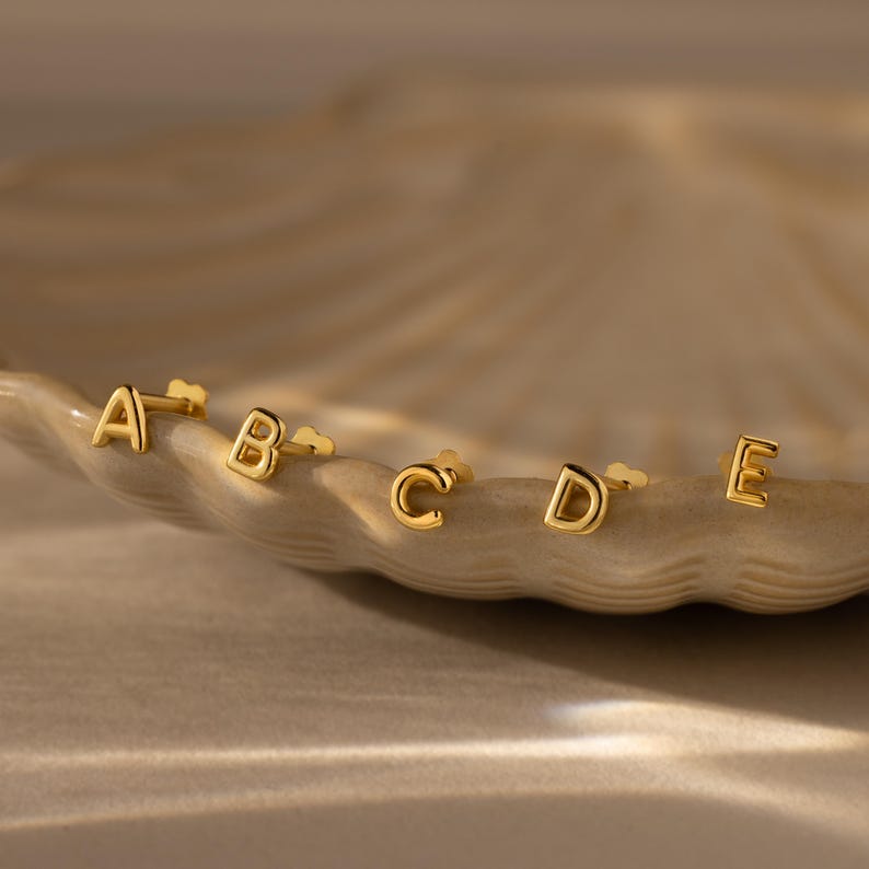 Gold initial stud earrings displaying the letters A, B, C, D, and E, arranged along the edge of a textured dish under warm, soft lighting.