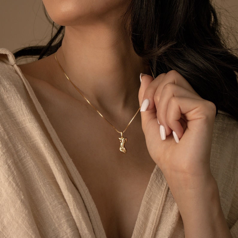 May include: A gold chain necklace with a pendant shaped like a stylized human figure.