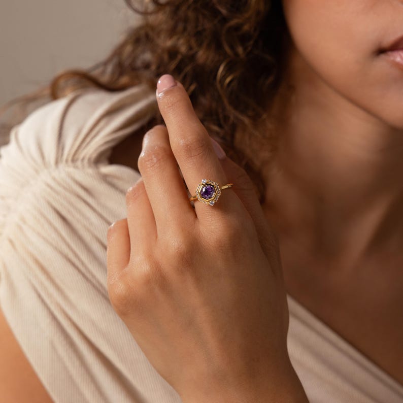 May include: A gold ring featuring a central round purple gemstone set within a hexagonal frame. The hexagonal setting is accented with small, clear, sparkling stones along its edges, mounted on a thin, polished gold band.