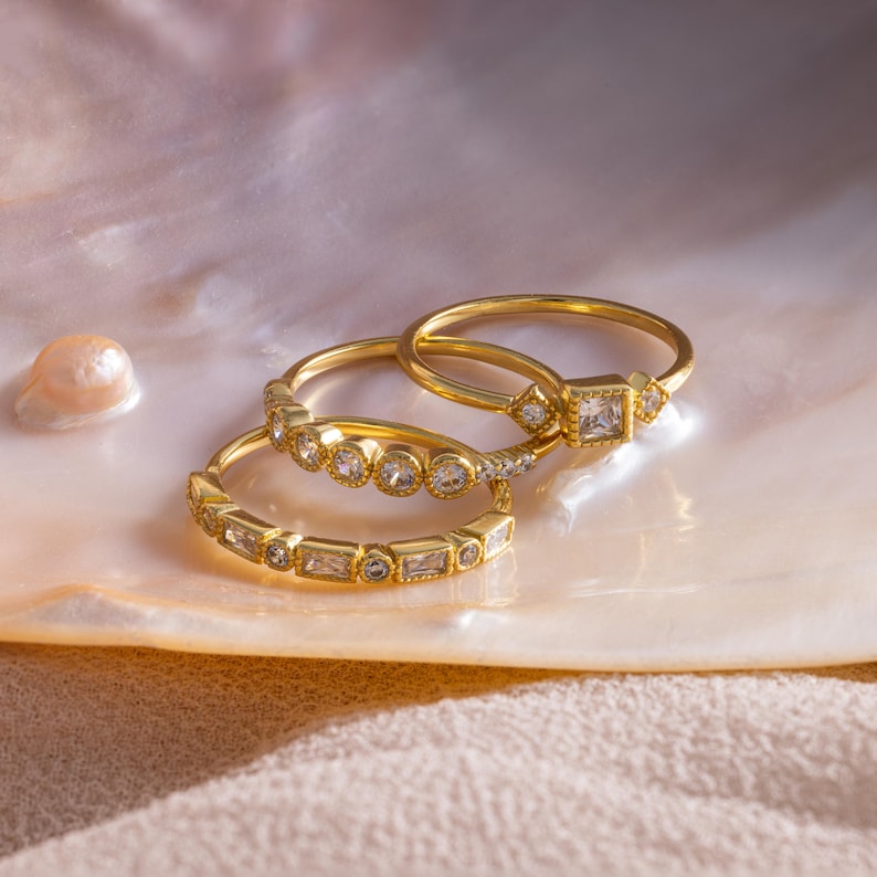 May include: Three gold rings with clear gemstones are displayed on a pearlescent surface. The rings feature various gemstone cuts, including round, square, and baguette shapes. The rings are set in gold bands, creating a luxurious and elegant look.