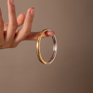 Pode incluir: Uma pulseira rígida articulada de dois tons com uma metade em dourado polido e uma metade em prateado polido. A pulseira circular tem uma superfície metálica lisa e reflexiva com um mecanismo de dobradiça visível que une as duas secções.