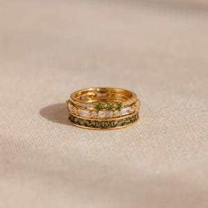 May include: A stack of four gold rings. The top ring has small clear stones. The second ring has clear and green stones. The third ring has clear stones. The bottom ring has green stones. The rings are on a neutral background.