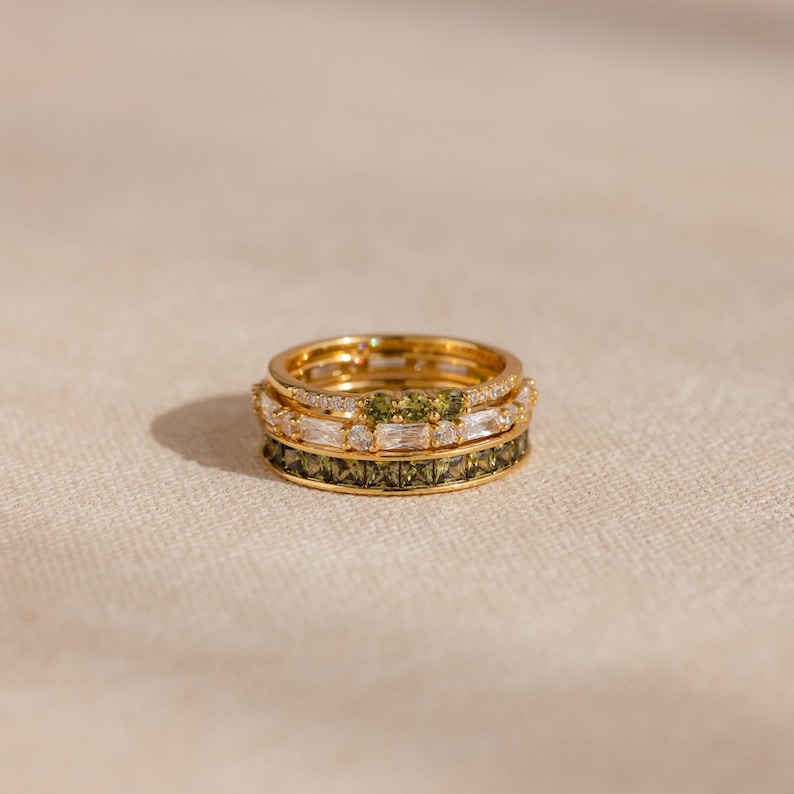 May include: A stack of four gold rings. The top ring has small clear stones. The second ring has clear and green stones. The third ring has clear stones. The bottom ring has green stones. The rings are on a neutral background.