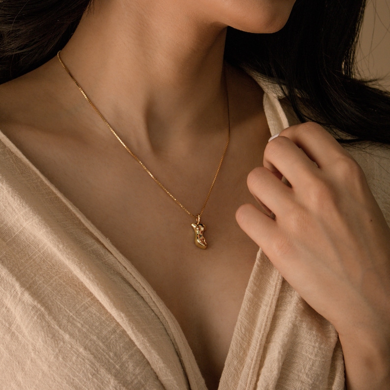 May include: A gold chain necklace with a small, abstract, gold pendant. The pendant is shaped like a human figure.