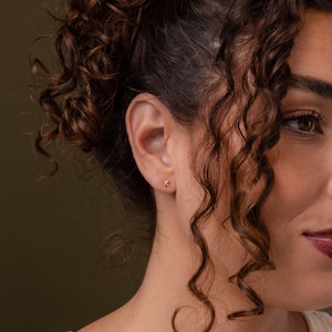 Close-up of a woman with curly hair wearing a small gold letter K stud earring, styled in warm natural lighting with soft shadows.