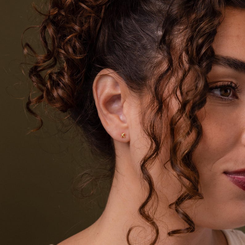 Close-up of a woman with curly hair wearing a small gold letter K stud earring, styled in warm natural lighting with soft shadows.