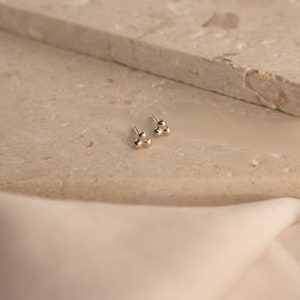 May include: A pair of silver stud earrings with three small, round balls clustered together. The earrings are on a light beige surface.