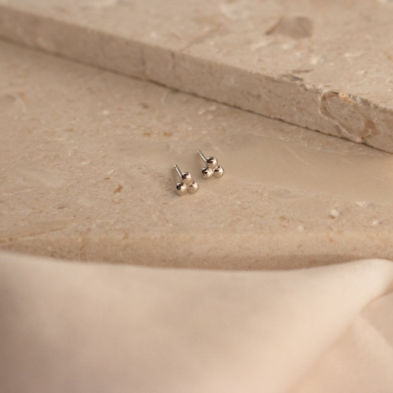 May include: A pair of silver stud earrings with three small, round balls clustered together. The earrings are on a light beige surface.