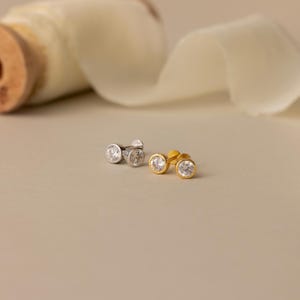 May include: Two stud earrings featuring round, clear, faceted stones set in smooth metal bezels. One earring is finished in a bright silver tone, while the other is finished in a warm gold tone, both showcasing a clean, circular design.