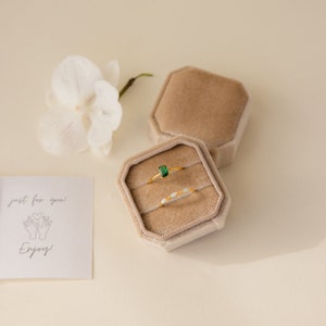 May include: Two gold rings in a beige velvet ring box. The top ring has a green emerald stone. The bottom ring has small white stones. The box is open and the rings are visible. The text "just for you!" and a drawing of two hands holding a heart is on a white card.