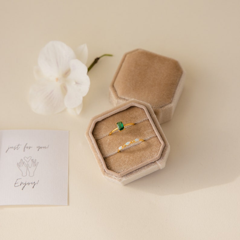 May include: Two gold rings in a beige velvet ring box. The top ring has a green emerald stone. The bottom ring has small white stones. The box is open and the rings are visible. The text "just for you!" and a drawing of two hands holding a heart is on a white card.