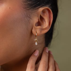 May include: A gold hoop earring with a small bow and a teardrop-shaped gemstone pendant. The earring is worn on the ear. The background is a soft green color.