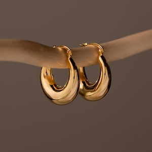 Pode incluir: Um par de brincos de argola em ouro polido com um design espesso, arredondado e cónico. Os brincos apresentam uma superfície metálica lisa e refletora com um mecanismo de fecho de dobradiça, exibindo uma forma curva contínua e brilhante.
