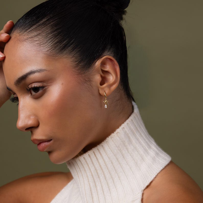 May include: A close-up of a gold hoop earring with a small bow and a teardrop-shaped clear gemstone. The earring is worn on a person's ear. The person is wearing a white turtleneck sweater.