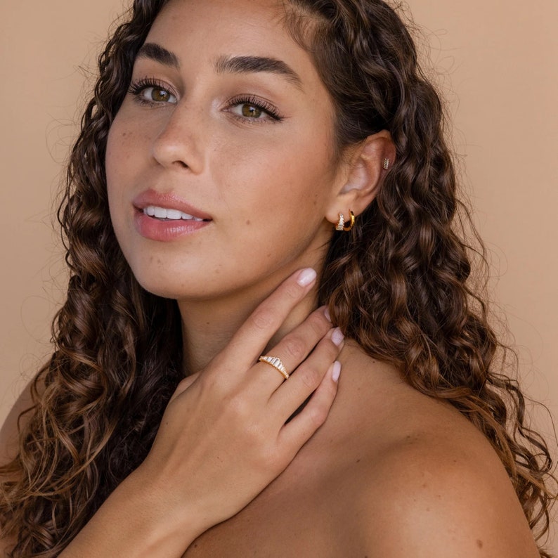 May include: A woman with long, curly brown hair is wearing a gold ring with small clear stones on her ring finger. She is wearing gold hoop earrings in one ear.