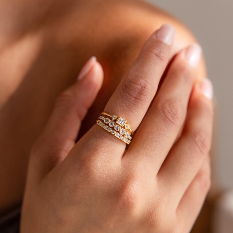 May include: A hand displays three gold rings with various gemstone settings. The rings feature round, square, and baguette-cut stones. The gold bands have a textured appearance, and the gemstones sparkle against the warm lighting.
