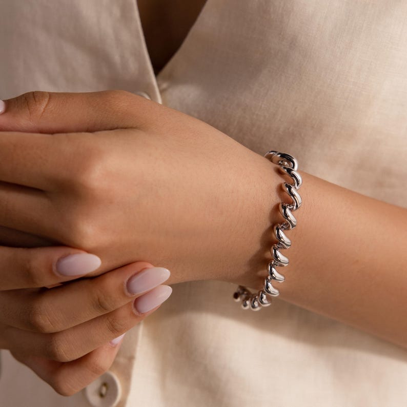 May include: A silver bracelet with a spiral design is worn on a wrist. The bracelet is made of shiny metal and has a series of curved links. The skin tone is light, and the nails are painted with a light pink colour.