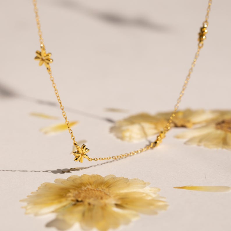 May include: A delicate gold chain necklace with small flower charms. The necklace is photographed on a white background with dried flowers.
