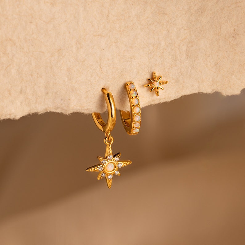 May include: A set of three gold earrings featuring a starburst design with opal accents. The earrings are on a beige background.