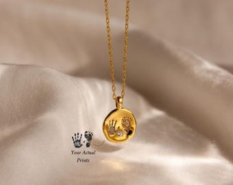 May include: A gold-toned necklace featuring a circular pendant with an engraved handprint and footprint design. The pendant is accented with a small, clear, round-cut gemstone set between the prints. The chain is a delicate gold-toned link style.
