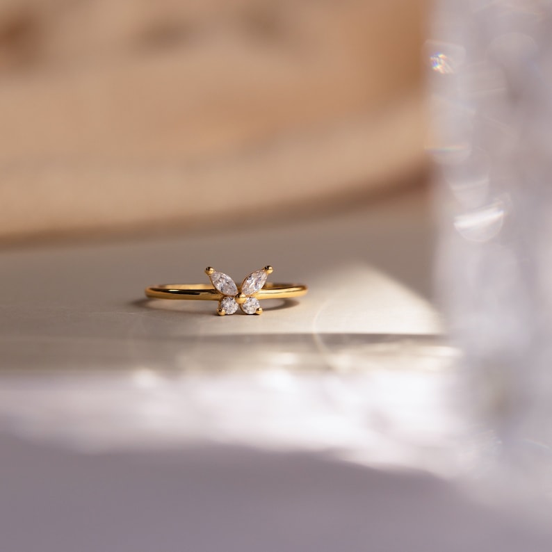 A close up of our Mariposa Butterfly Ring in 18K Gold finish placed on a white table and a clear glass vase in the foreground - featuring an about 5.5mm x 8mm CZ Diamond Butterfly Charm and an about 1.5mm Thin Ring Band.