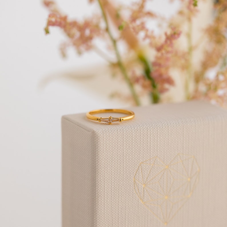 A close up of our Eleanor Diamond Ring in 18K Gold finish set on one of our jewelry gift boxes with pink flowers in the background - featuring a 1.5mm ring band and a 9mm CZ Diamond Baguette Stone.