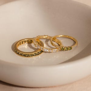May include: Three gold rings with different gemstone designs. One ring features olive-green stones, another has clear baguette-cut stones, and the third has small olive-green stones. The rings are displayed in a white bowl.