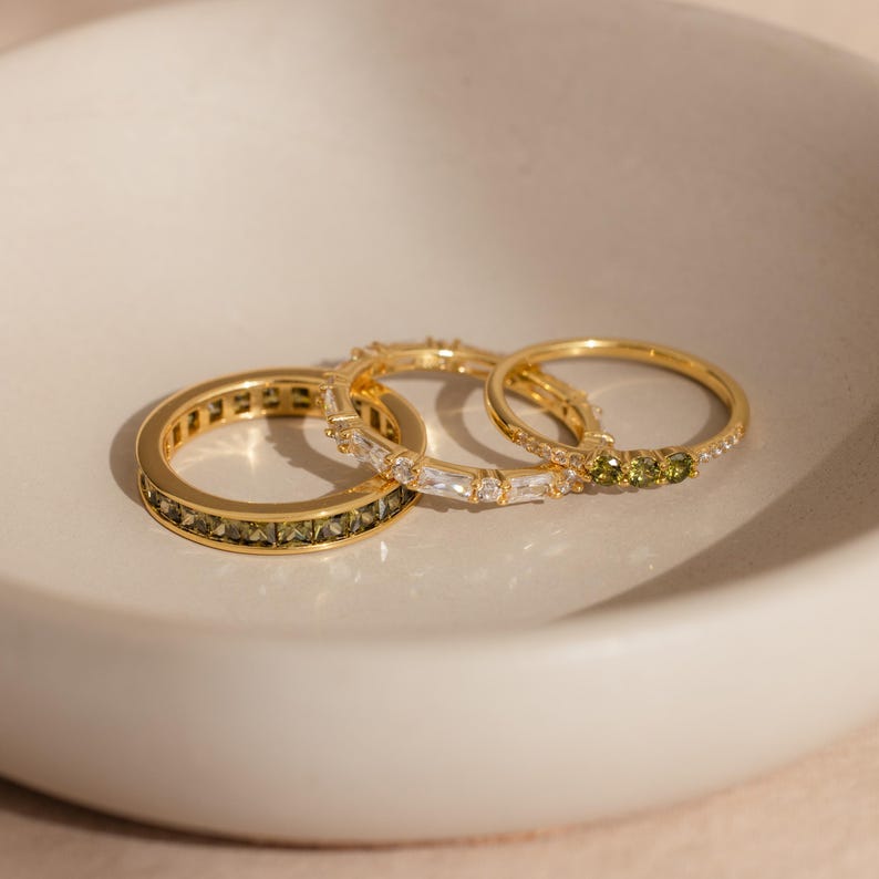 May include: Three gold rings with different gemstone designs. One ring features olive-green stones, another has clear baguette-cut stones, and the third has small olive-green stones. The rings are displayed in a white bowl.