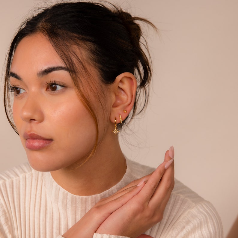 May include: A woman wearing gold earrings with a starburst design, a small hoop earring, and a tiny stud earring.