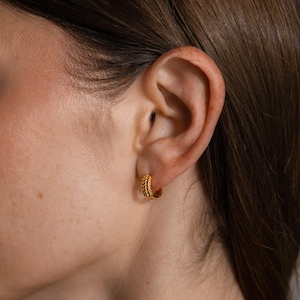 May include: Gold-colored hoop earrings with a textured, beaded design. The earrings are small and round, with a polished finish. The image shows the earrings worn on an ear, highlighting their size and style. These earrings are a classic jewelry accessory.