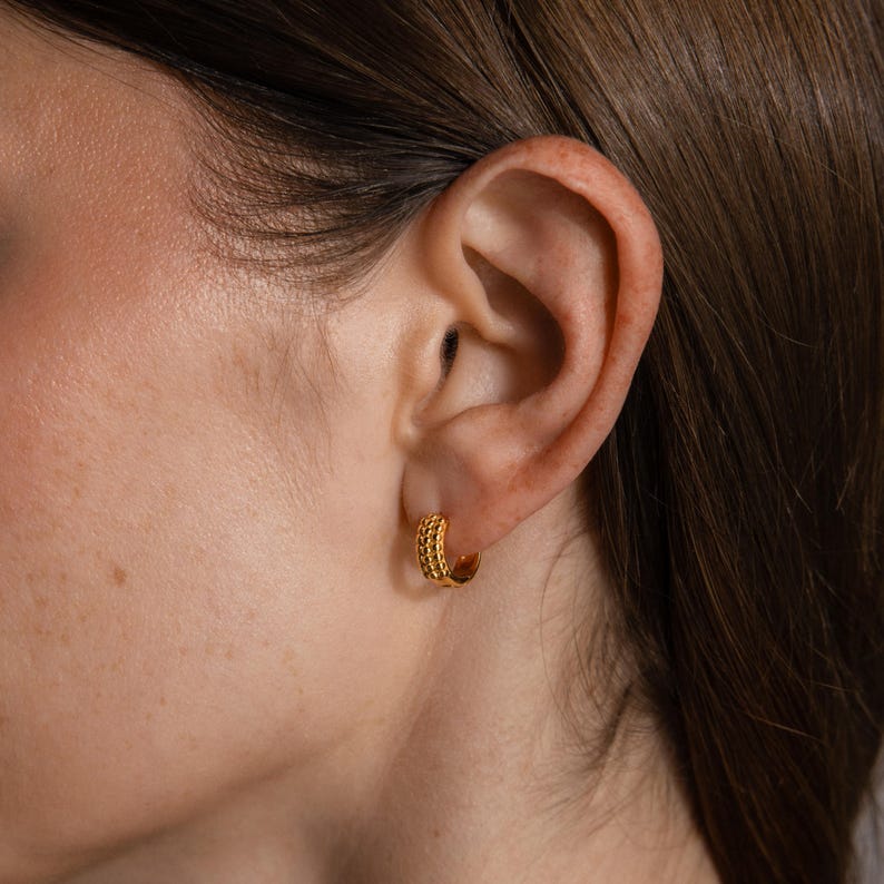 May include: Gold-colored hoop earrings with a textured, beaded design. The earrings are small and round, with a polished finish. The image shows the earrings worn on an ear, highlighting their size and style. These earrings are a classic jewelry accessory.