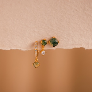 May include: A set of gold earrings with green and clear stones. The earrings are on a light brown background.