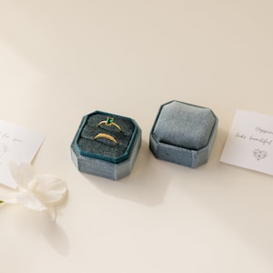 May include: Two small, square, blue velvet ring boxes. One box is open and contains three gold rings, one with a green emerald stone. The other box is closed. The text on the card reads "Happiness looks beautiful".