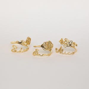 May include: Three gold-colored metal rings, each featuring a delicate floral design attached to a script name. The names displayed in cursive lettering are Sadie, Emma, and Alice. Each ring has a thin band and a decorative flower element.