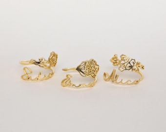 May include: Three gold-colored metal rings, each featuring a delicate floral design attached to a script name. The names displayed in cursive lettering are Sadie, Emma, and Alice. Each ring has a thin band and a decorative flower element.