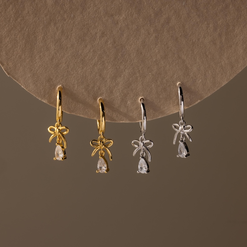 May include: Gold and silver-toned hoop earrings with a bow and teardrop-shaped charm. The earrings are displayed on a neutral-toned background. The earrings are a delicate jewelry item.
