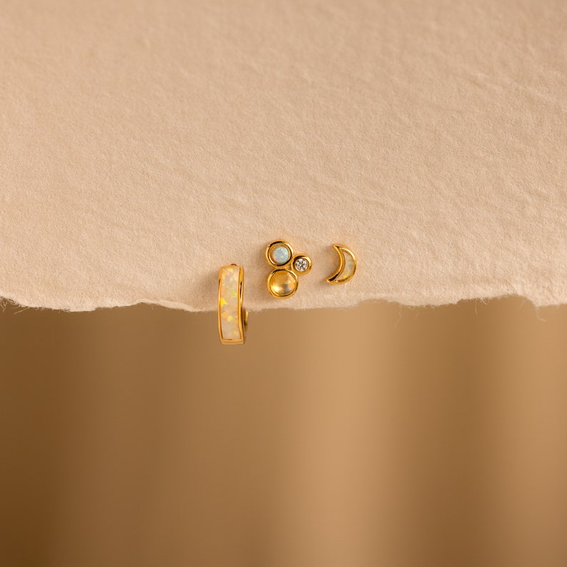 May include: Three gold earrings with opal and white stones. The earrings are a hoop, a crescent moon, and a cluster of three stones.