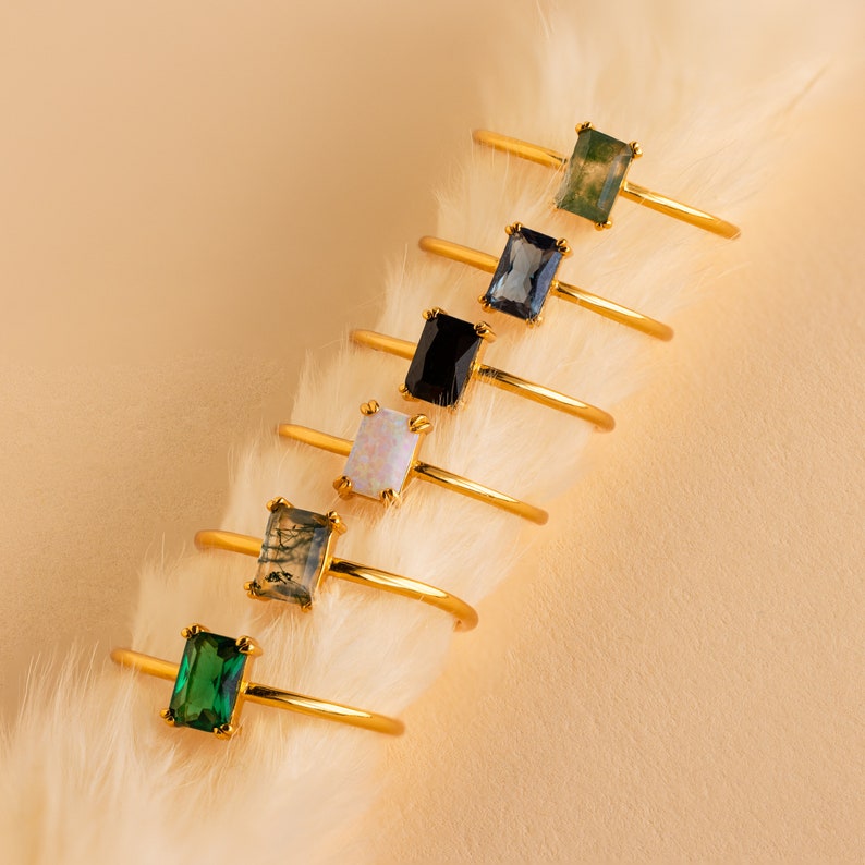 May include: A collection of gold rings with rectangular gemstones in various colors, including green, blue, black, and white. The rings are displayed on a white, fluffy surface against a beige background. The gemstones are set in a gold bezel.