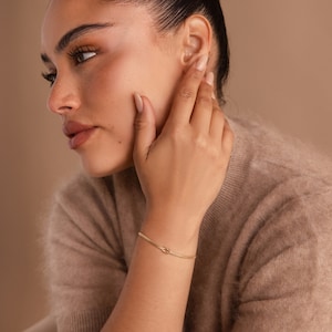 May include: A gold knot bracelet with a delicate chain design. The bracelet is worn on the wrist of a person with a tan complexion.