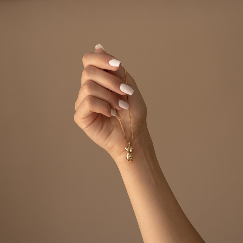 May include: A gold chain necklace with a small, detailed pendant in the shape of a stylized angel. The necklace is being held by a hand with white nail polish.
